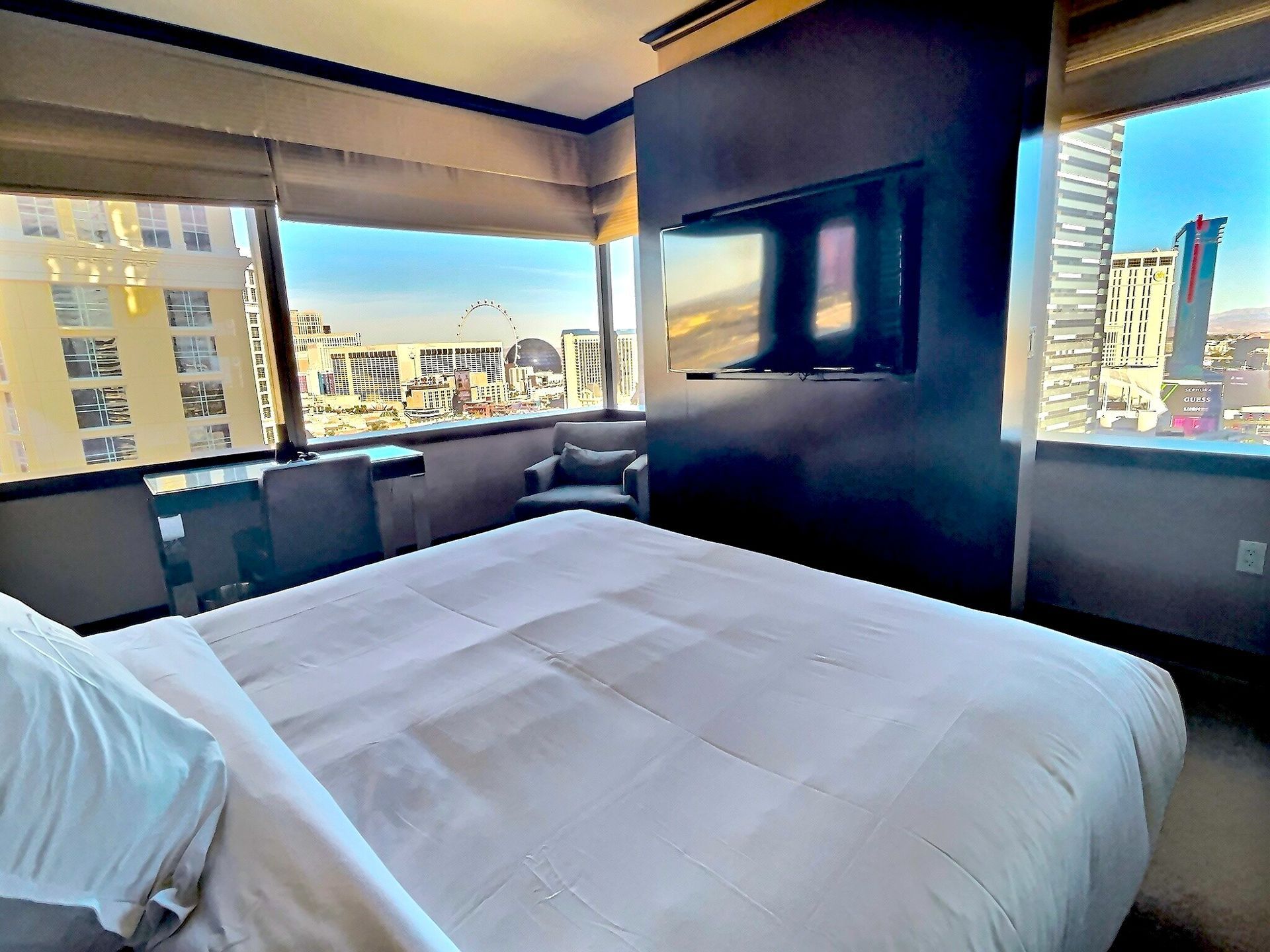 Modern hotel bedroom with a large bed, city-view windows, and a wall-mounted TV