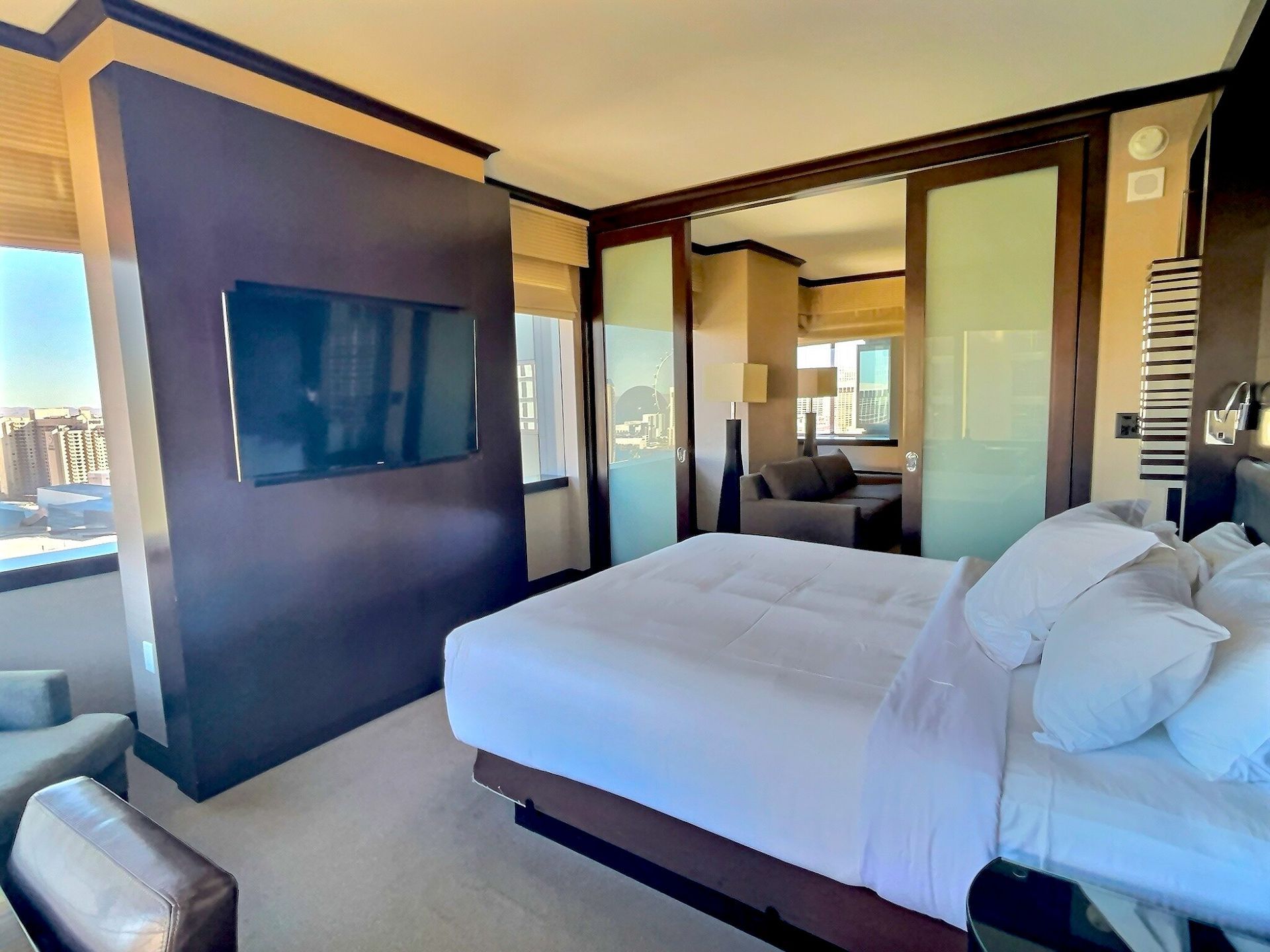 Modern hotel bedroom with king bed, wall-mounted TV, balcony doors, and city view