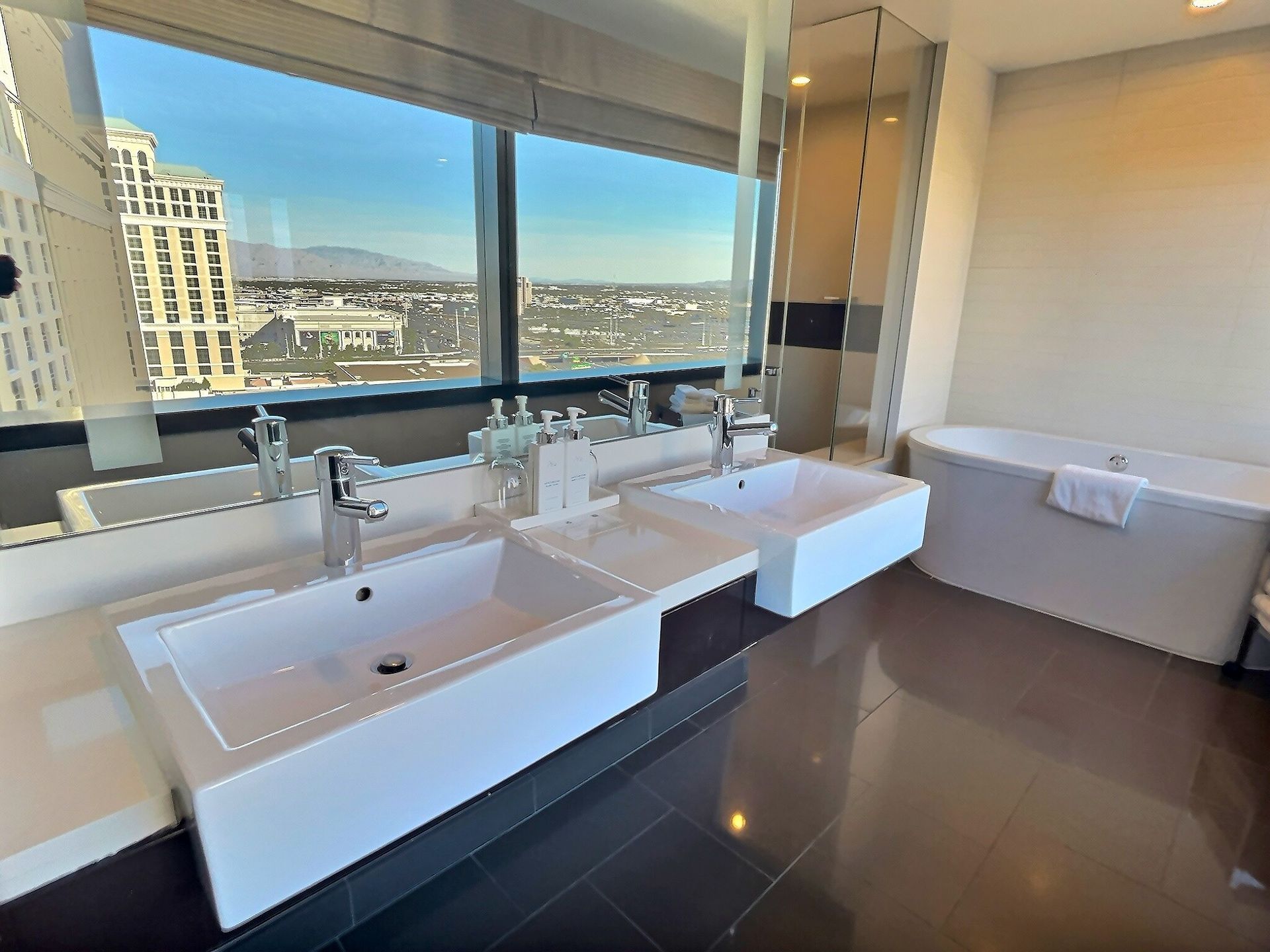 Modern bathroom with double sinks, large mirror, bathtub, and city view through wide windows
