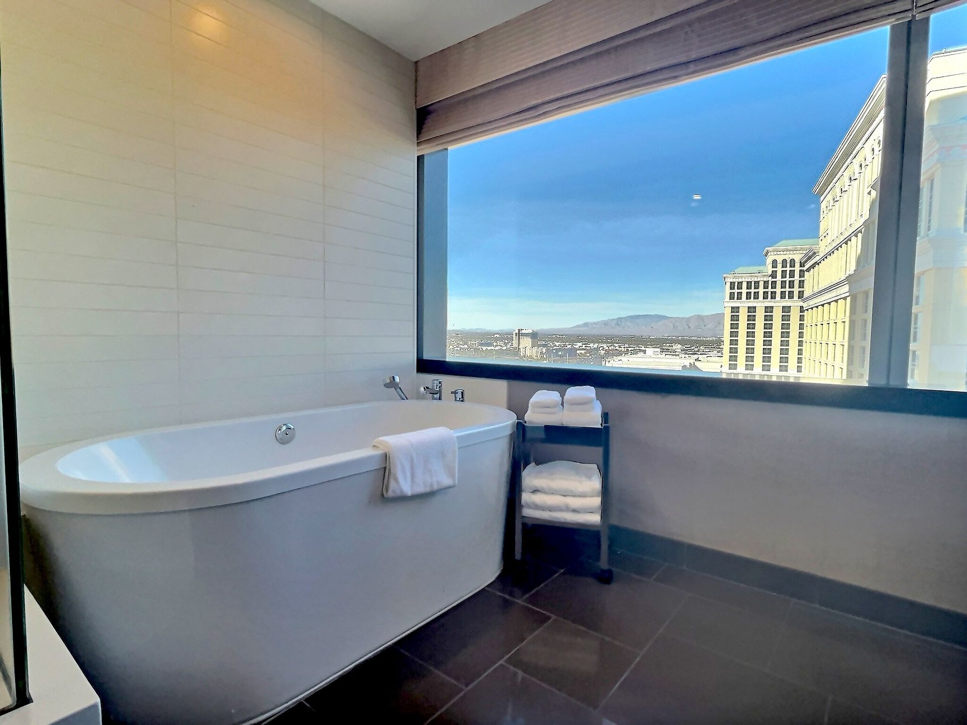 Modern bathroom with a soaking tub, folded towels, and a large window overlooking the city and mountains.