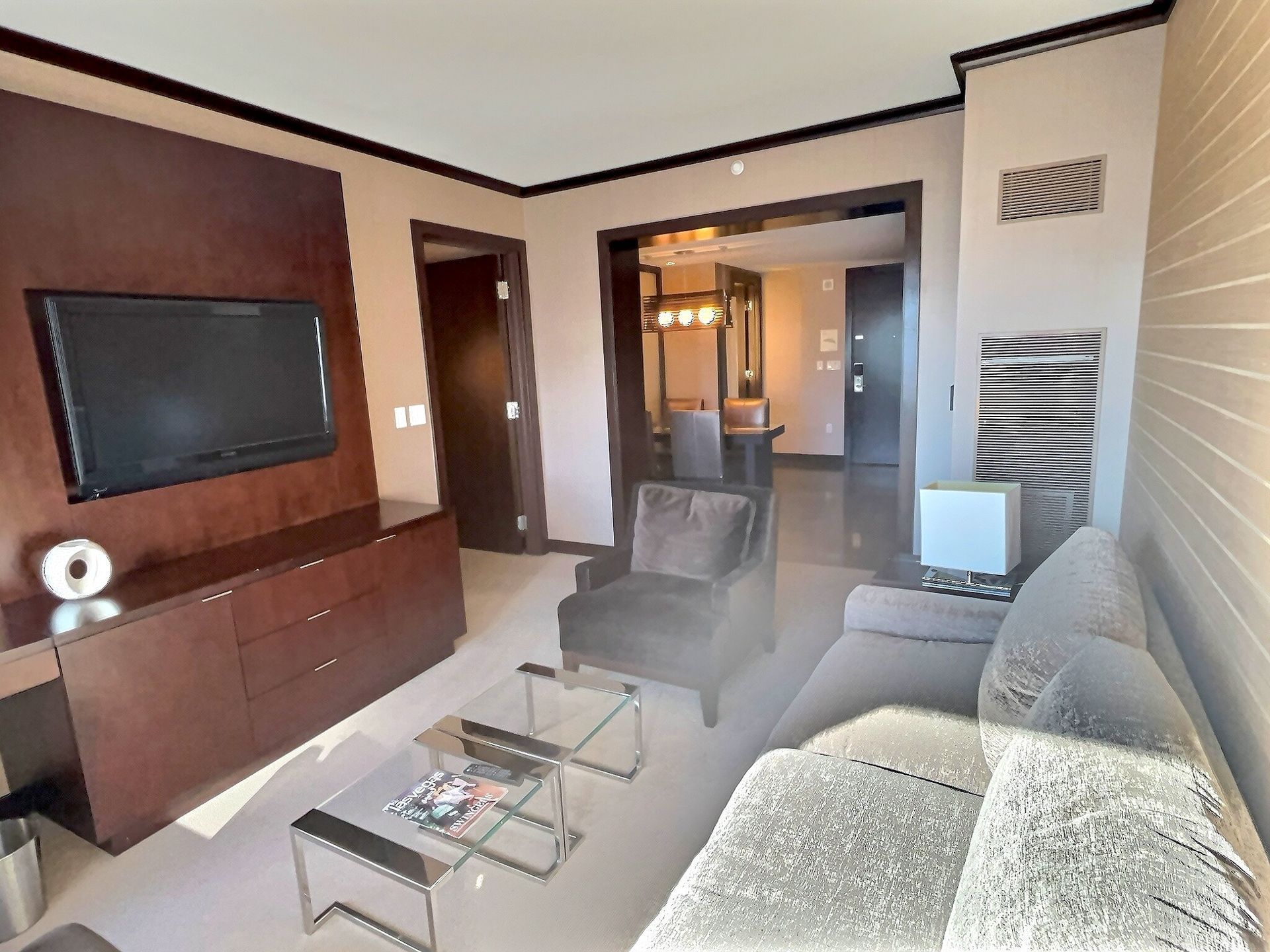 Modern living room with wall-mounted TV, glass coffee table, gray sofa, and open doorway to a hallway