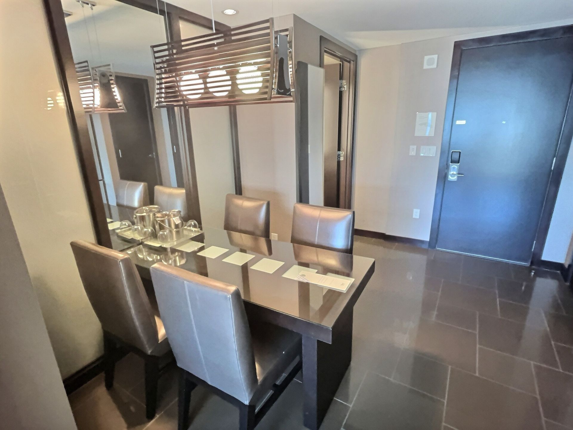 Modern dining room with glass table, four chairs, mirror wall, and dark entry door