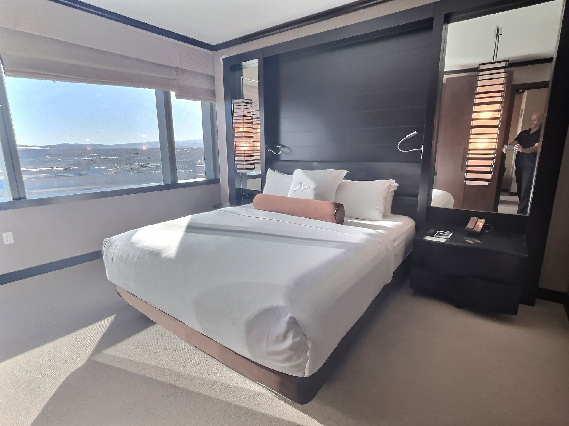 Modern hotel bedroom with king bed, dark accent wall, and large window overlooking the city.