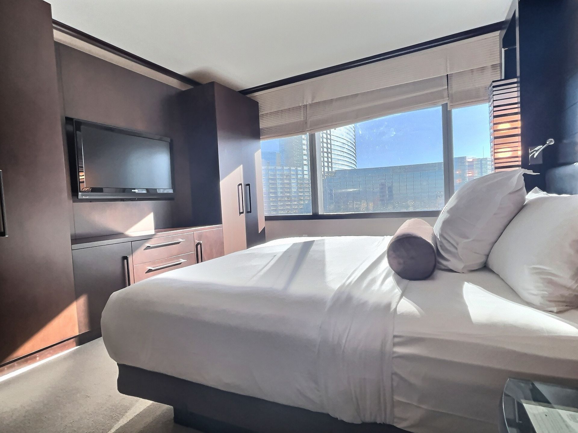 Modern hotel room with a bed, wall-mounted TV, and large window with city view
