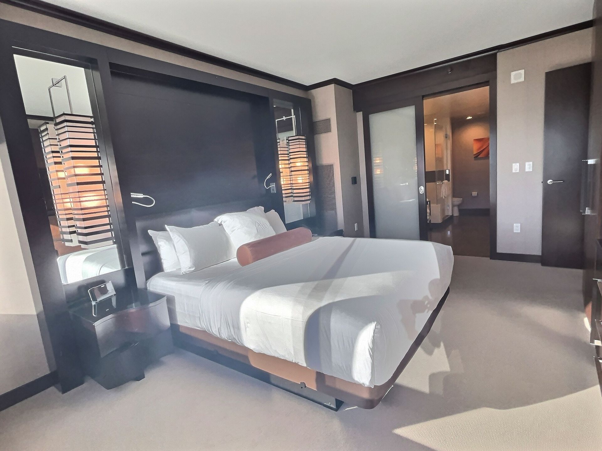 Modern hotel bedroom with a large bed, dark wood furnishings, and an en-suite bathroom door open.