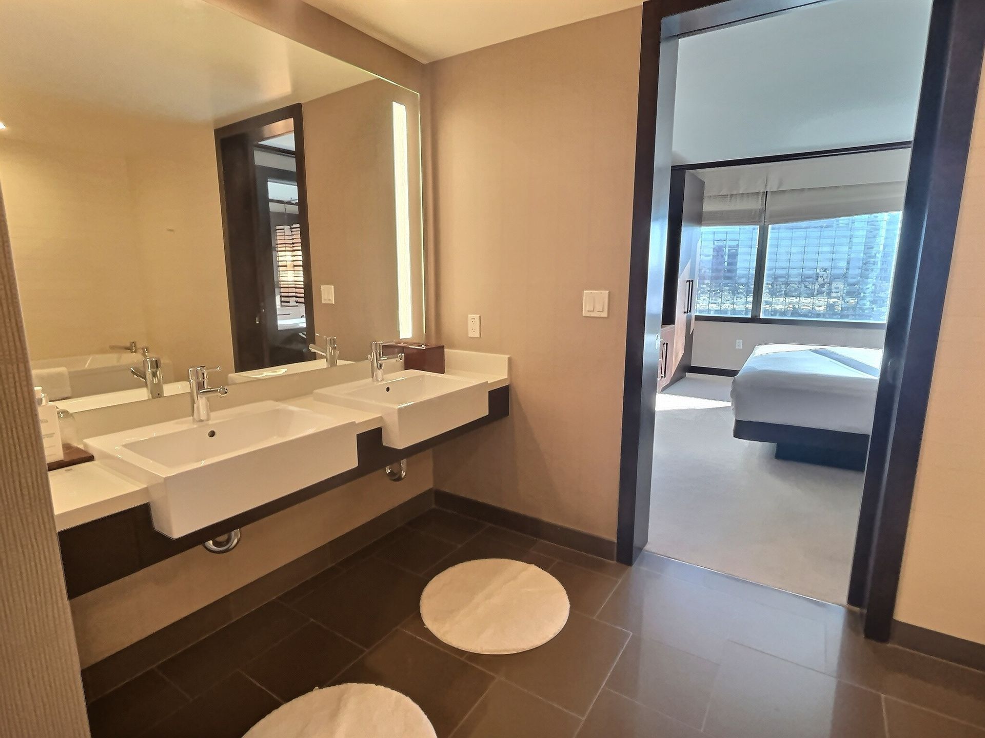Modern bathroom with double sink, large mirror, and view into a bedroom with a bed and city skyline.