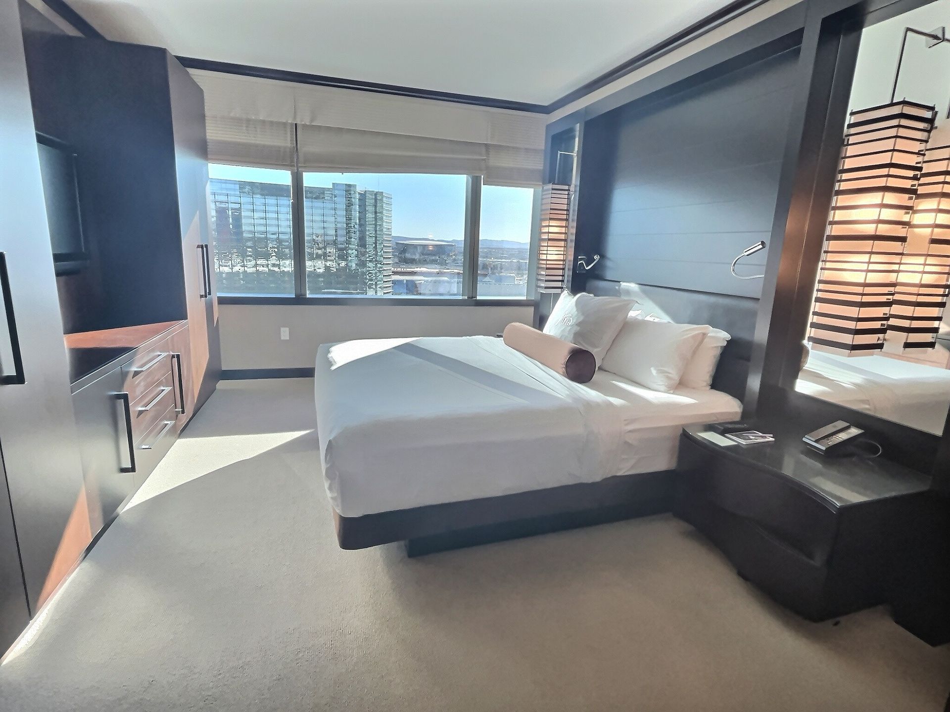 Modern bedroom with large windows, city view, white bed, gray carpet, and dark wood furniture.