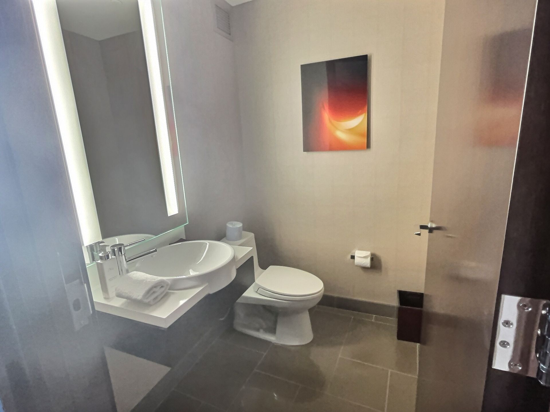 Public restroom with sink, toilet, mirror, gray tile floor, and wall art.