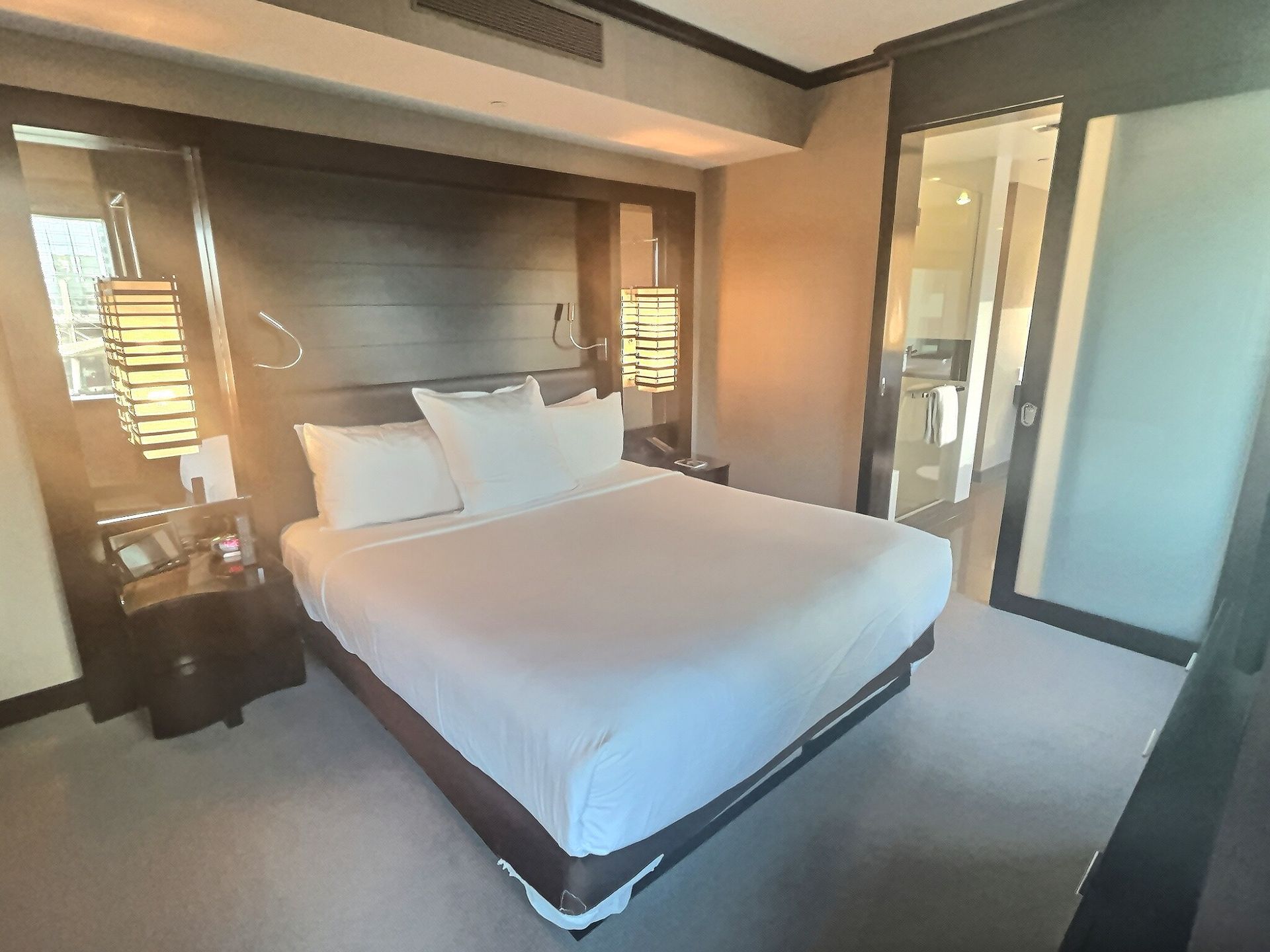 Modern hotel bedroom with a large bed, warm lighting, and glass doors to the bathroom