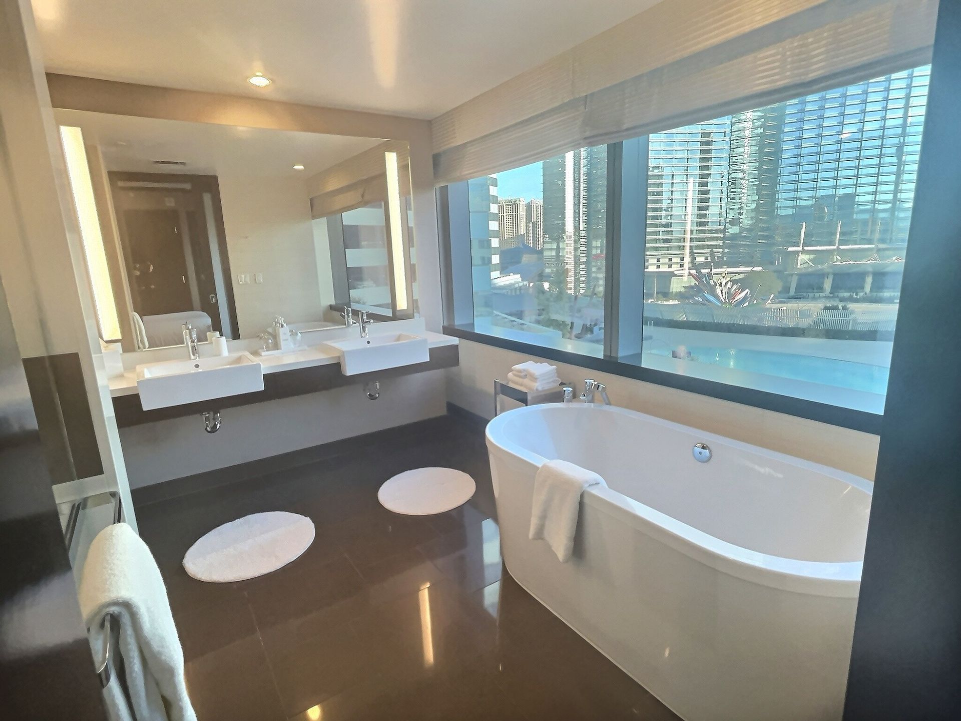 Modern bathroom with a large window, double sinks, round stools, and a freestanding tub overlooking a city view