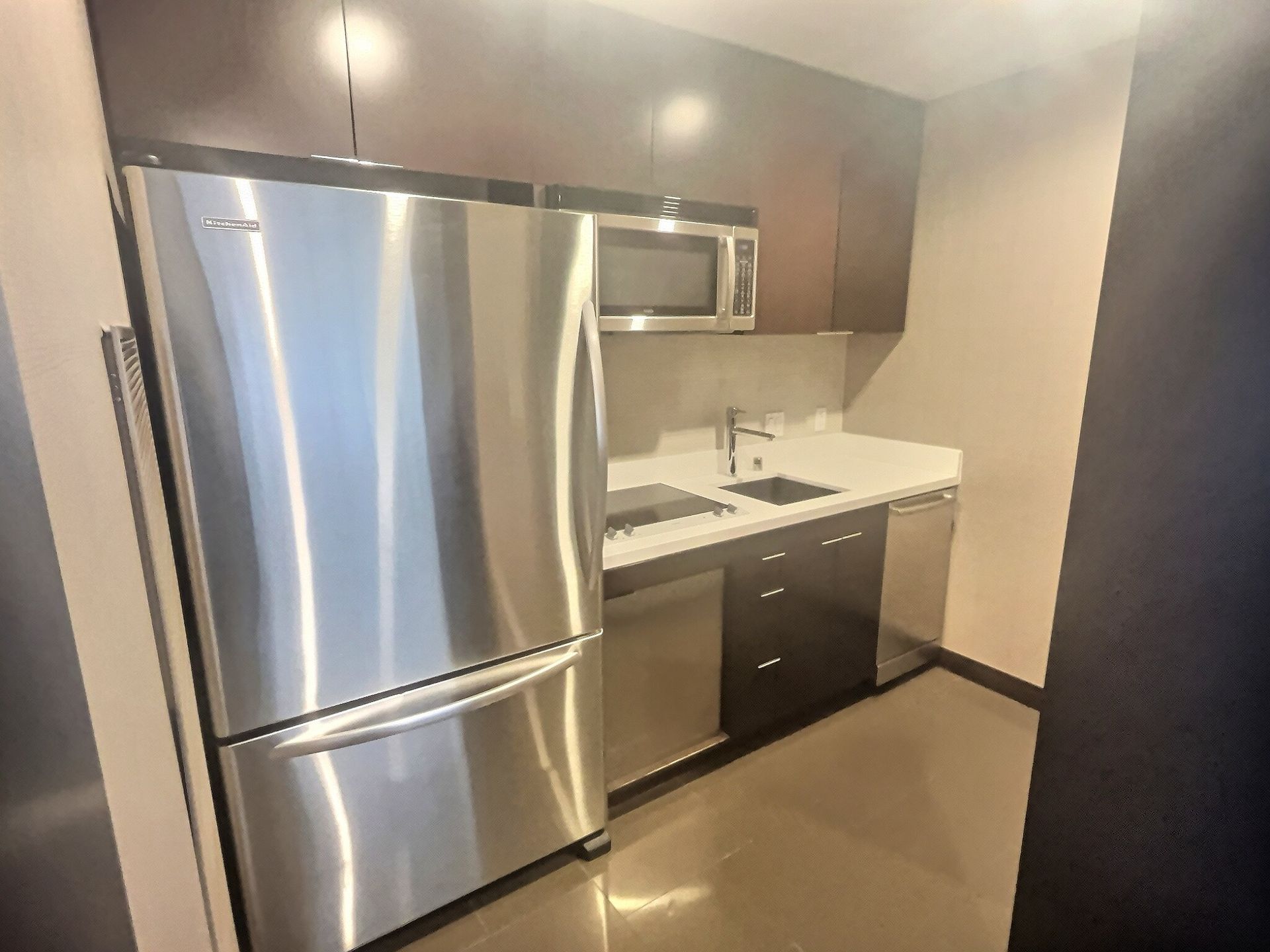 Compact kitchen with stainless steel fridge, microwave, sink, and dark cabinets.