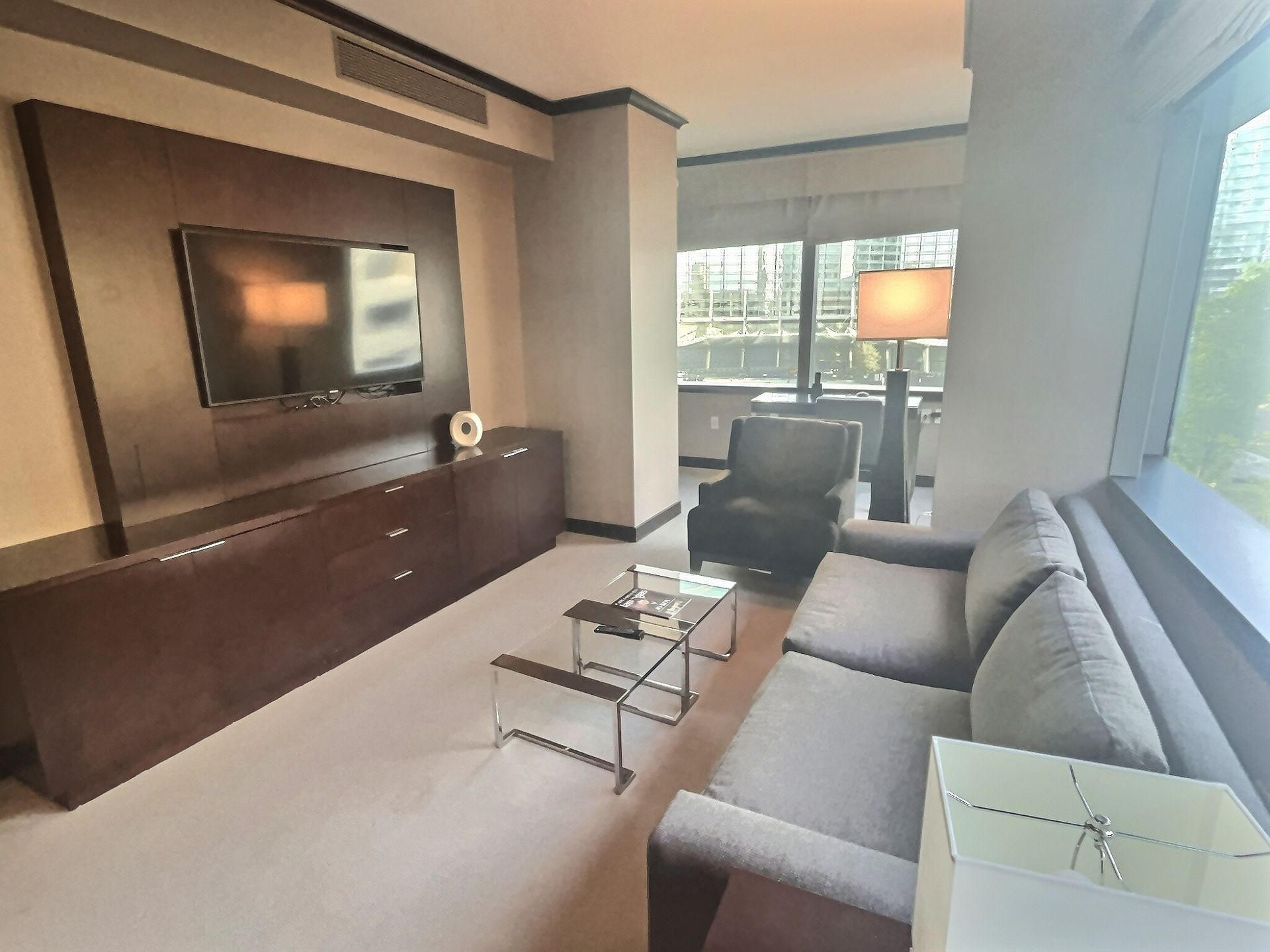 Modern living room with gray sectional sofa, wall-mounted TV, glass coffee table, and floor lamp by a window.