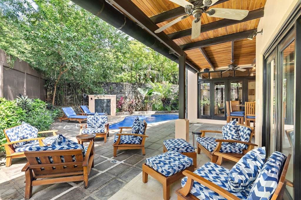 Covered patio with blue-cushioned wooden chairs and tables beside a backyard pool