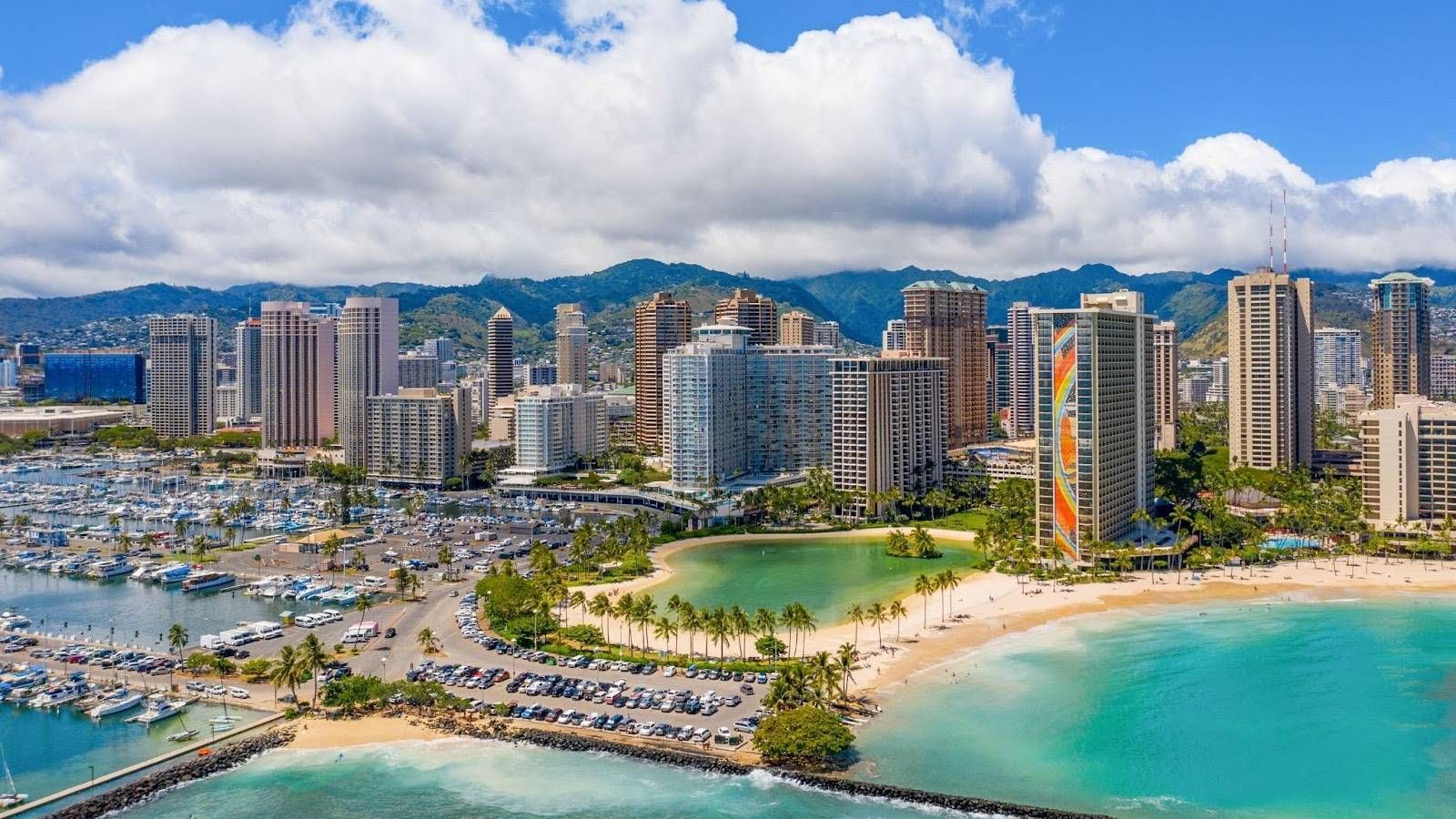 Aerial view of a beachfront city with high-rise hotels, marina boats, and turquoise water under cloudy skies
