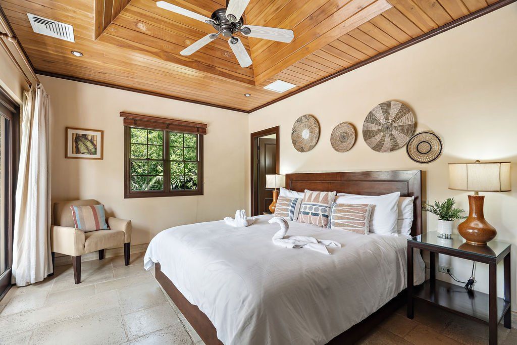 Bright bedroom with a white bed, wood ceiling, ceiling fan, and tropical decor in a sunlit home.