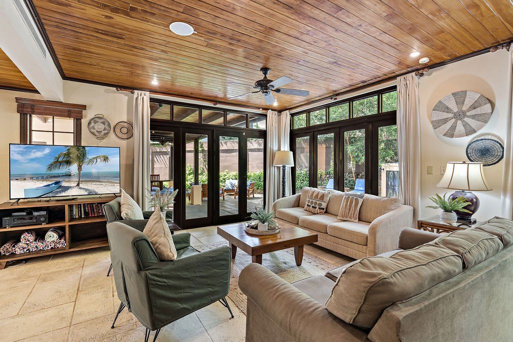 Bright sunroom living room with beige sofas, green chairs, large windows, and a wood-paneled ceiling