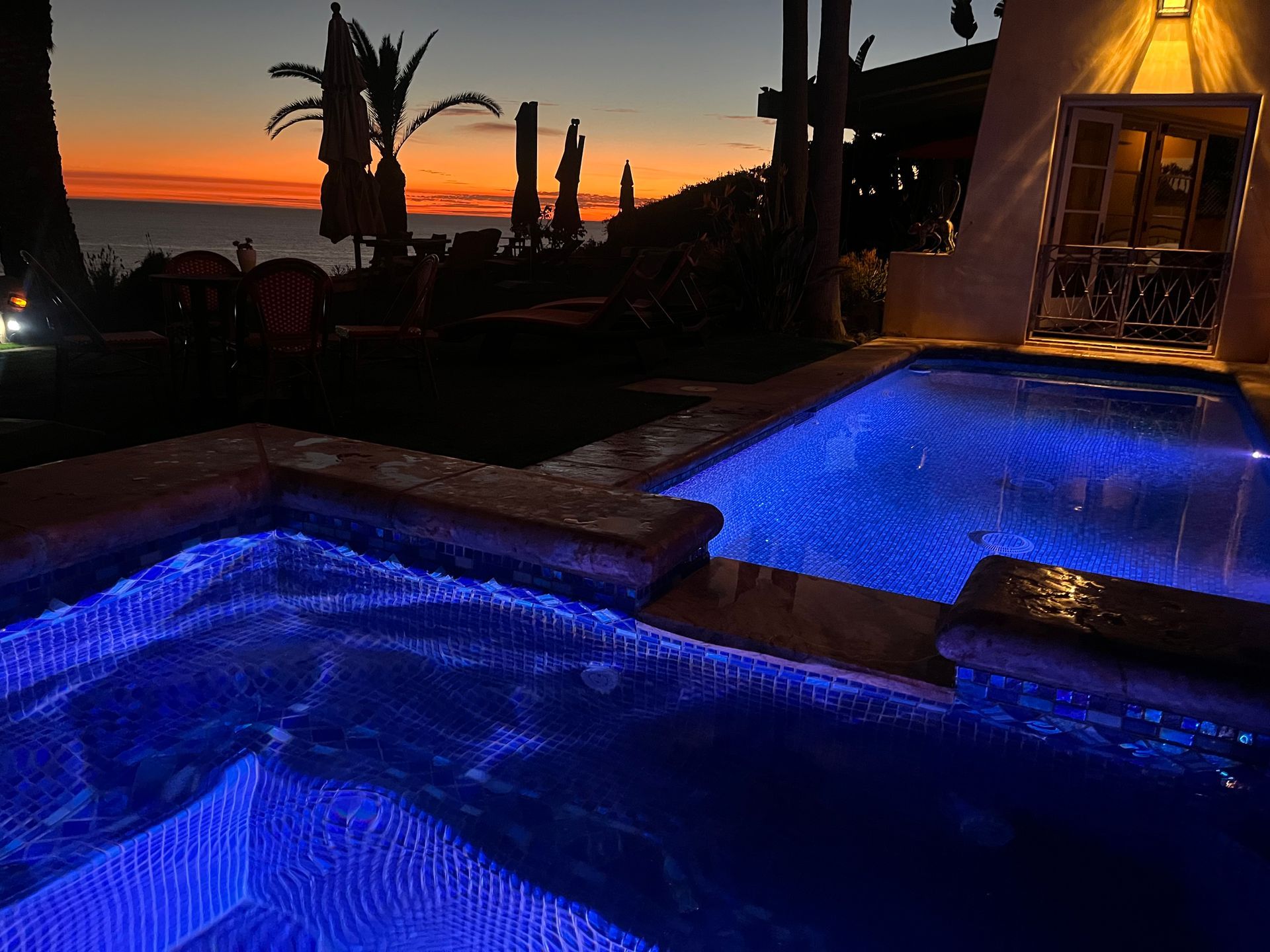 Blue-lit pool and spa at sunset with palm silhouettes and a glowing cabana window