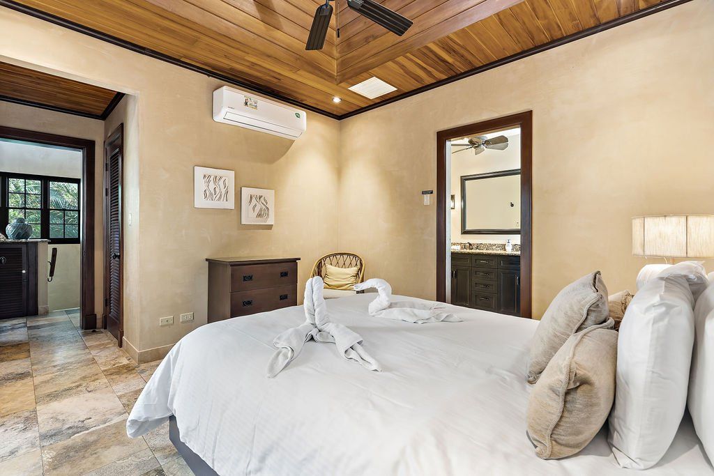 Bright bedroom with a large bed, wood ceiling, beige walls, and open doorway to a balcony.