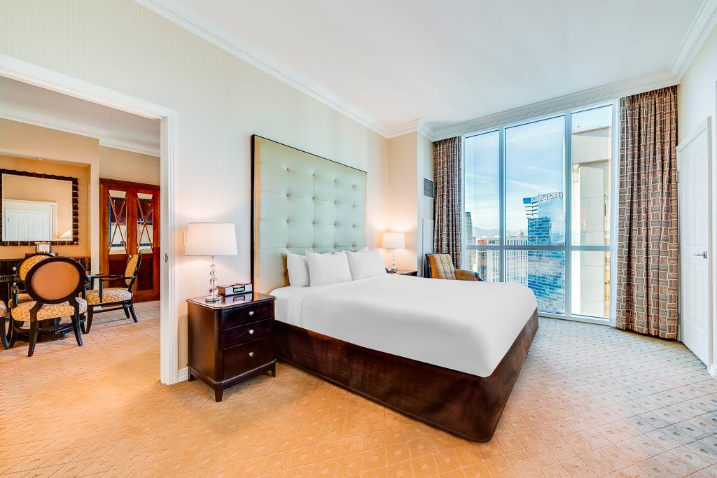 Bright hotel bedroom with king bed, city-view windows, and adjoining sitting area