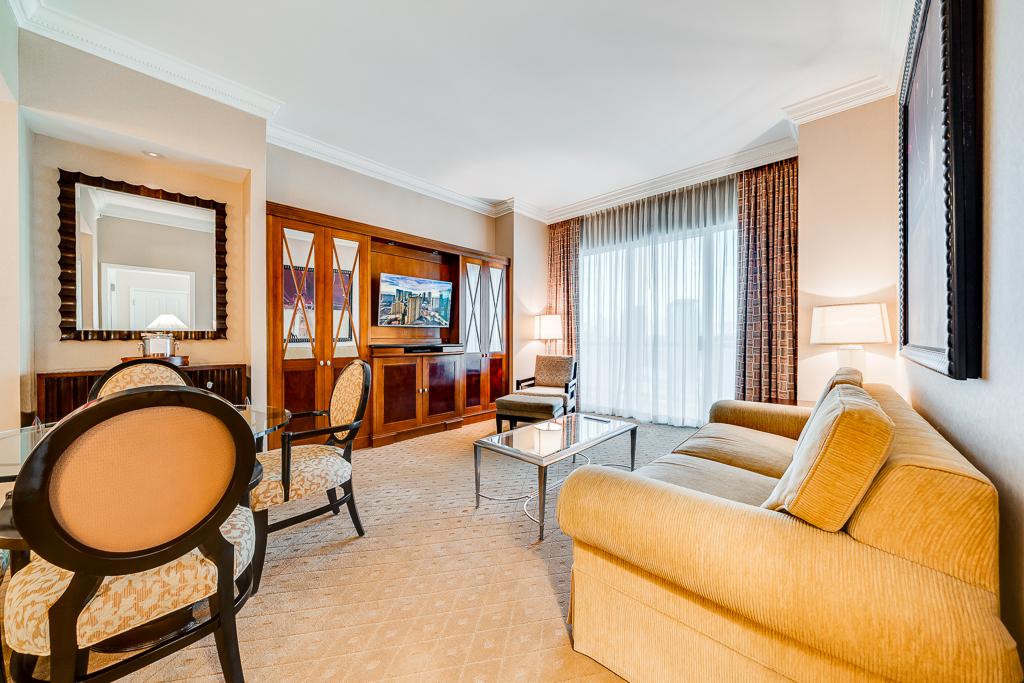 Elegant hotel living room with beige sofa, armchairs, glass coffee table, and large window with curtains