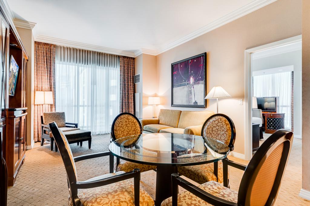 Elegant hotel room with glass dining table, chairs, desk, lamp, and city-view window with curtains