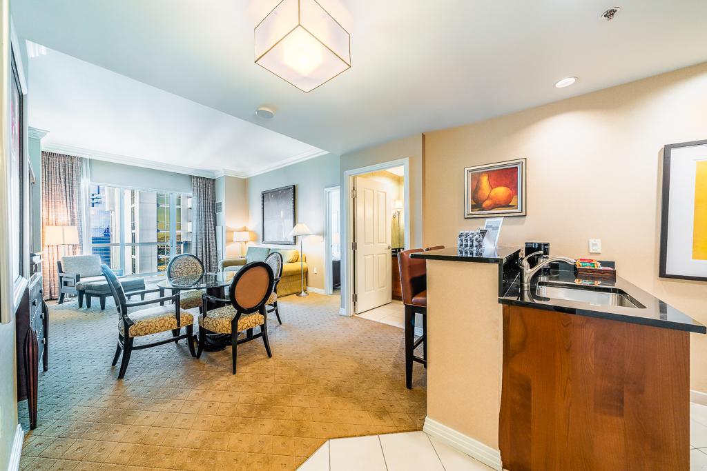 Bright hotel suite with living area, dining table, kitchenette, and beige carpet under soft lighting