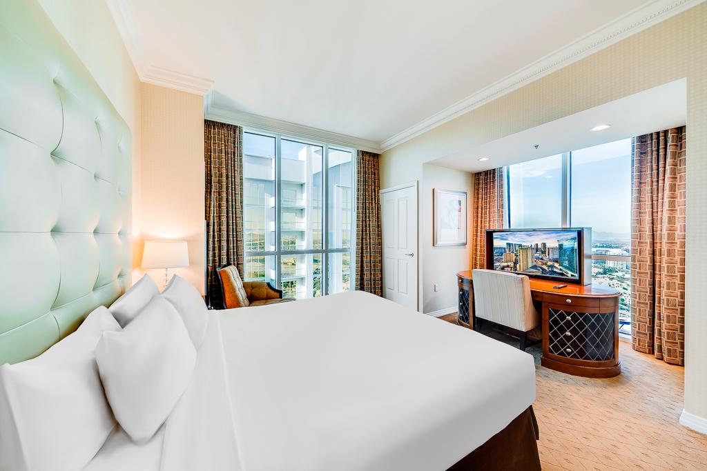 Bright hotel bedroom with a large bed, city-view windows, and a TV cabinet near the corner