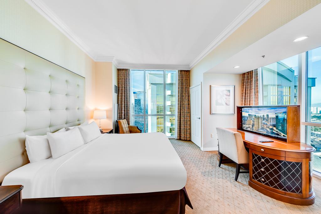 Bright hotel bedroom with king bed, tufted headboard, desk, TV, and balcony view