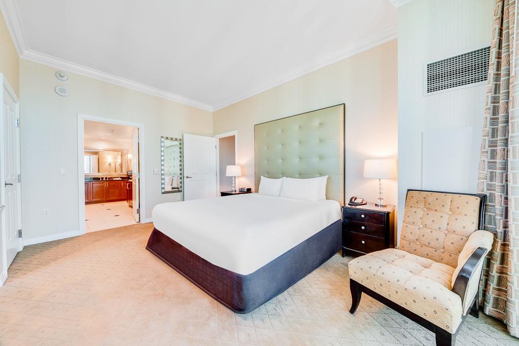 Bright bedroom with a large bed, tufted headboard, bedside lamp, and upholstered chair.