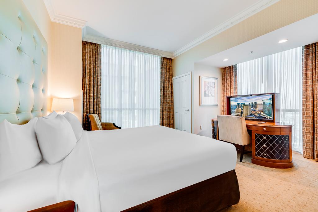Bright hotel room with a large white bed, desk, and city view through tall windows.