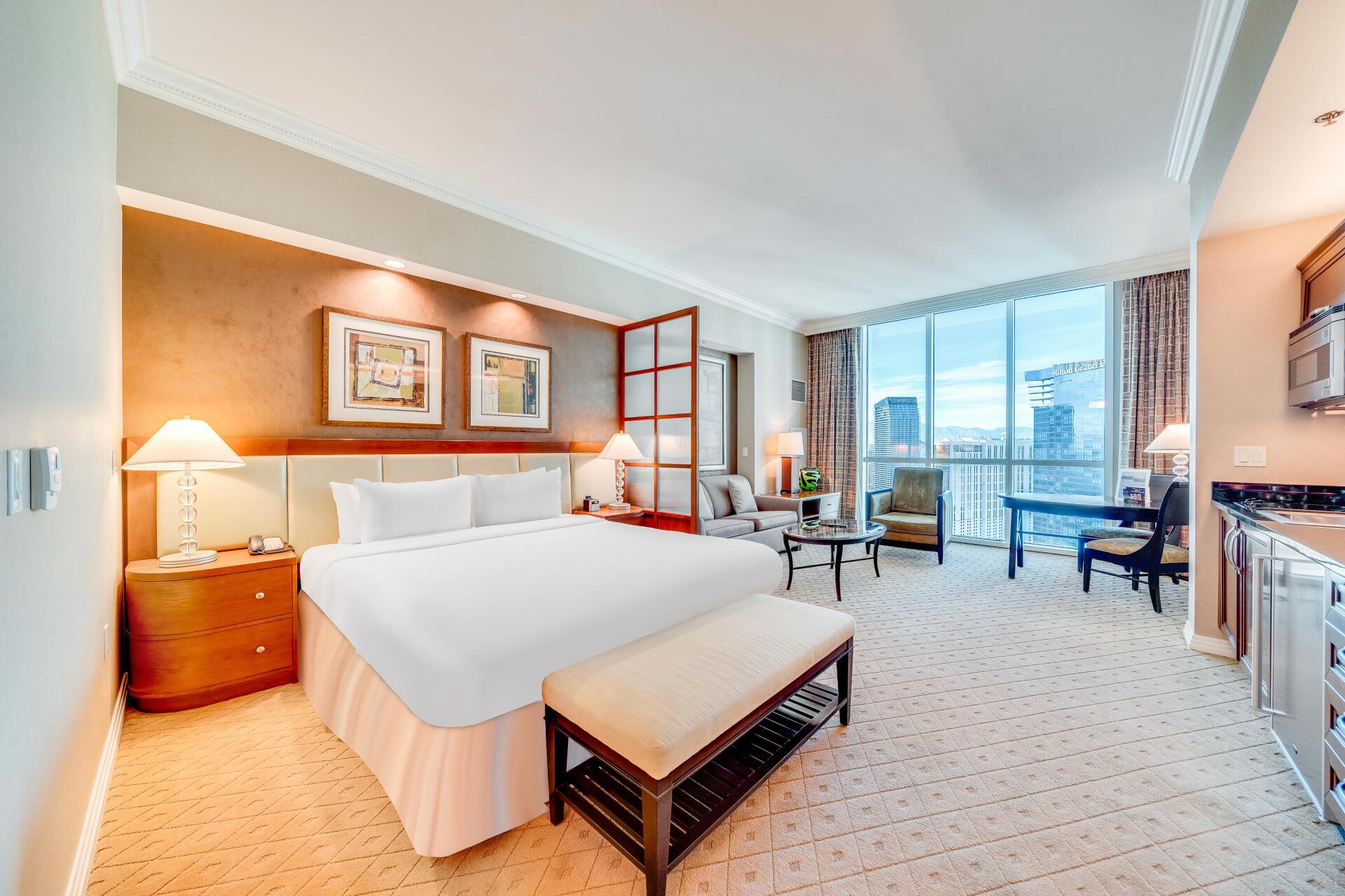 Spacious hotel room with king bed, sitting area, desk, and city view through large windows