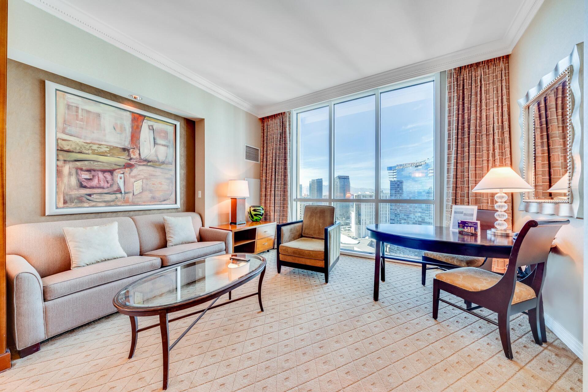 Bright living room with beige sofa, glass coffee table, armchairs, and city view through floor-to-ceiling windows