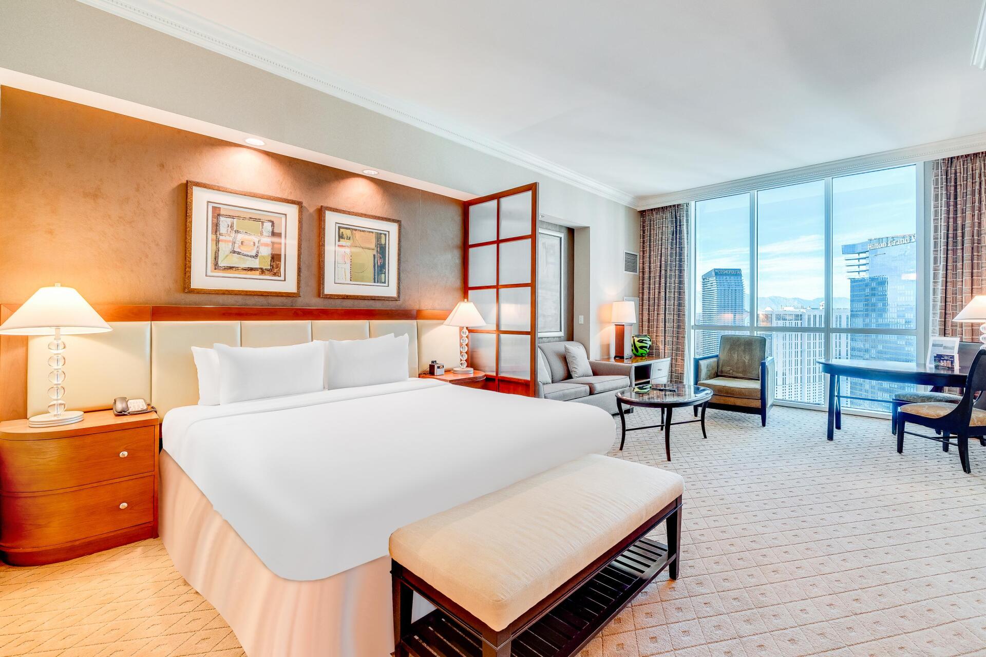 Bright hotel bedroom with a king bed, bench, desk, and city view through large windows.