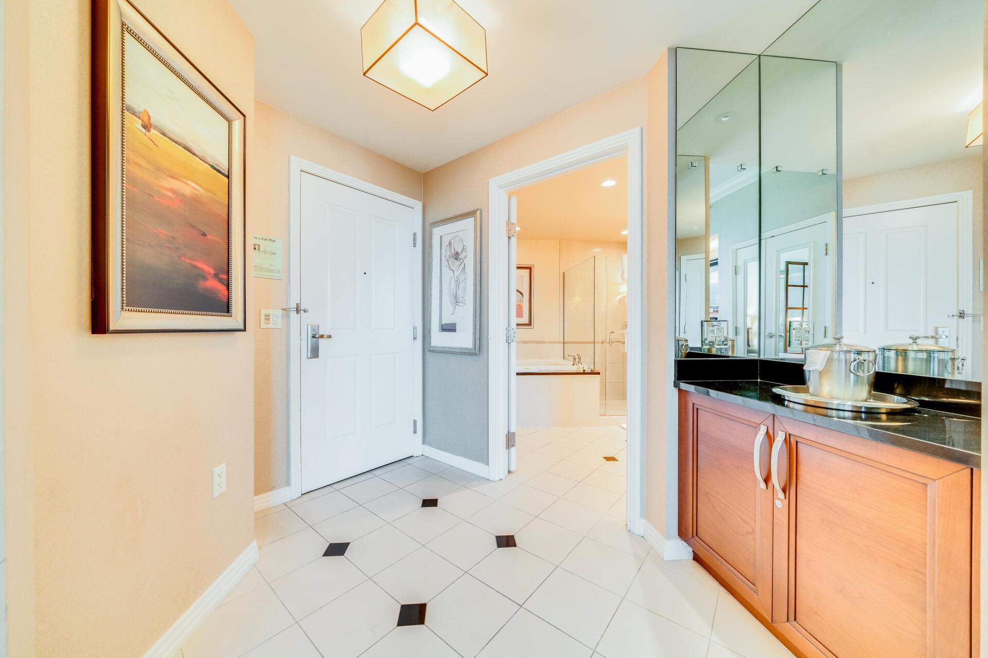 Bright hallway entryway with white tile floor, front door, framed art, and mirrored bathroom vanity counter.