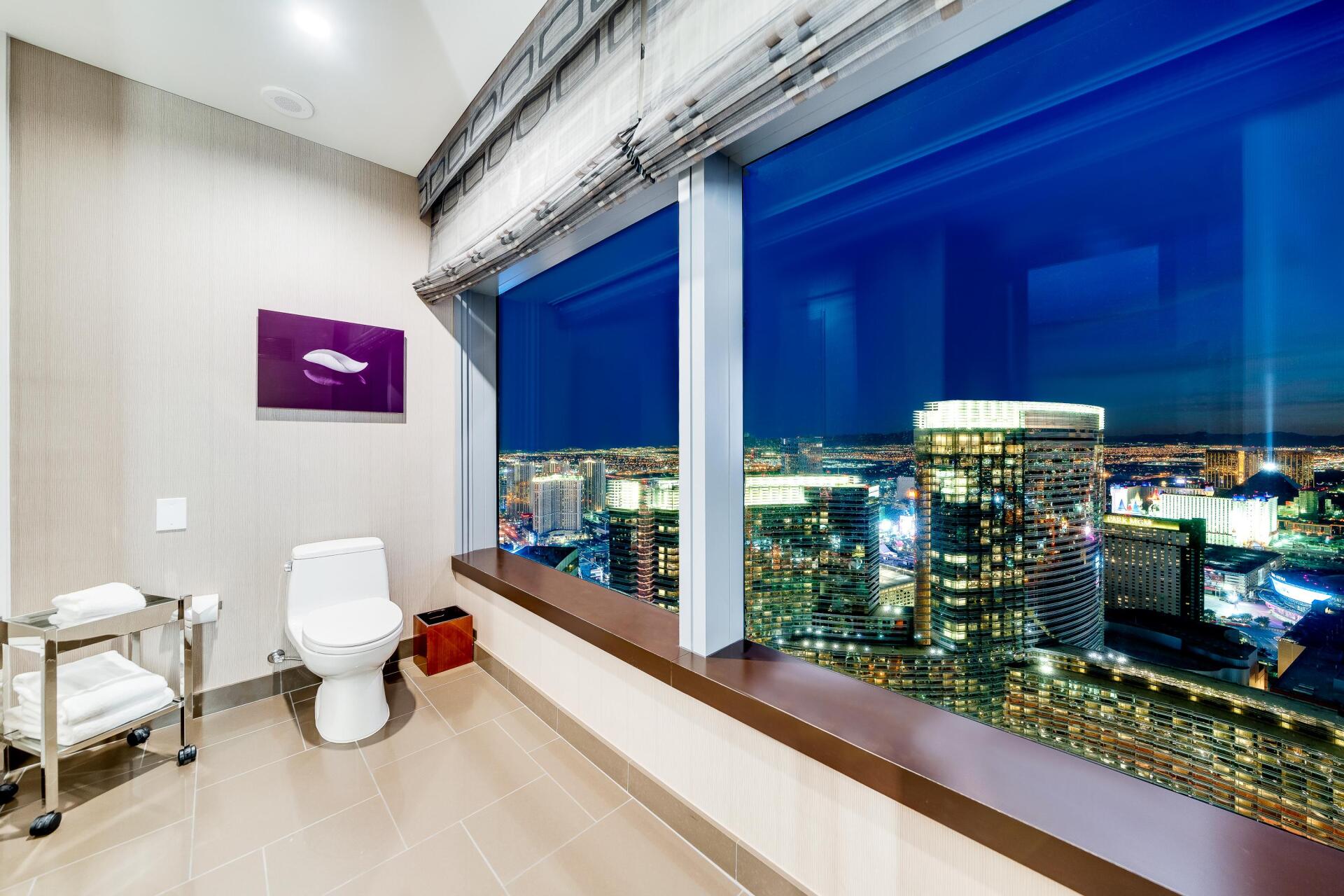 Modern bathroom with large city-view window at night and white toilet and chair