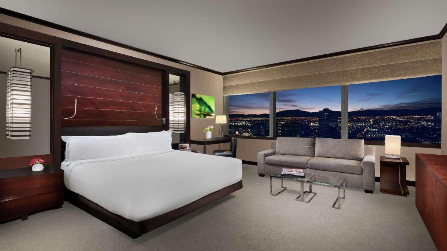 Modern hotel room with king bed, sofa, desk, and large city-view windows at night