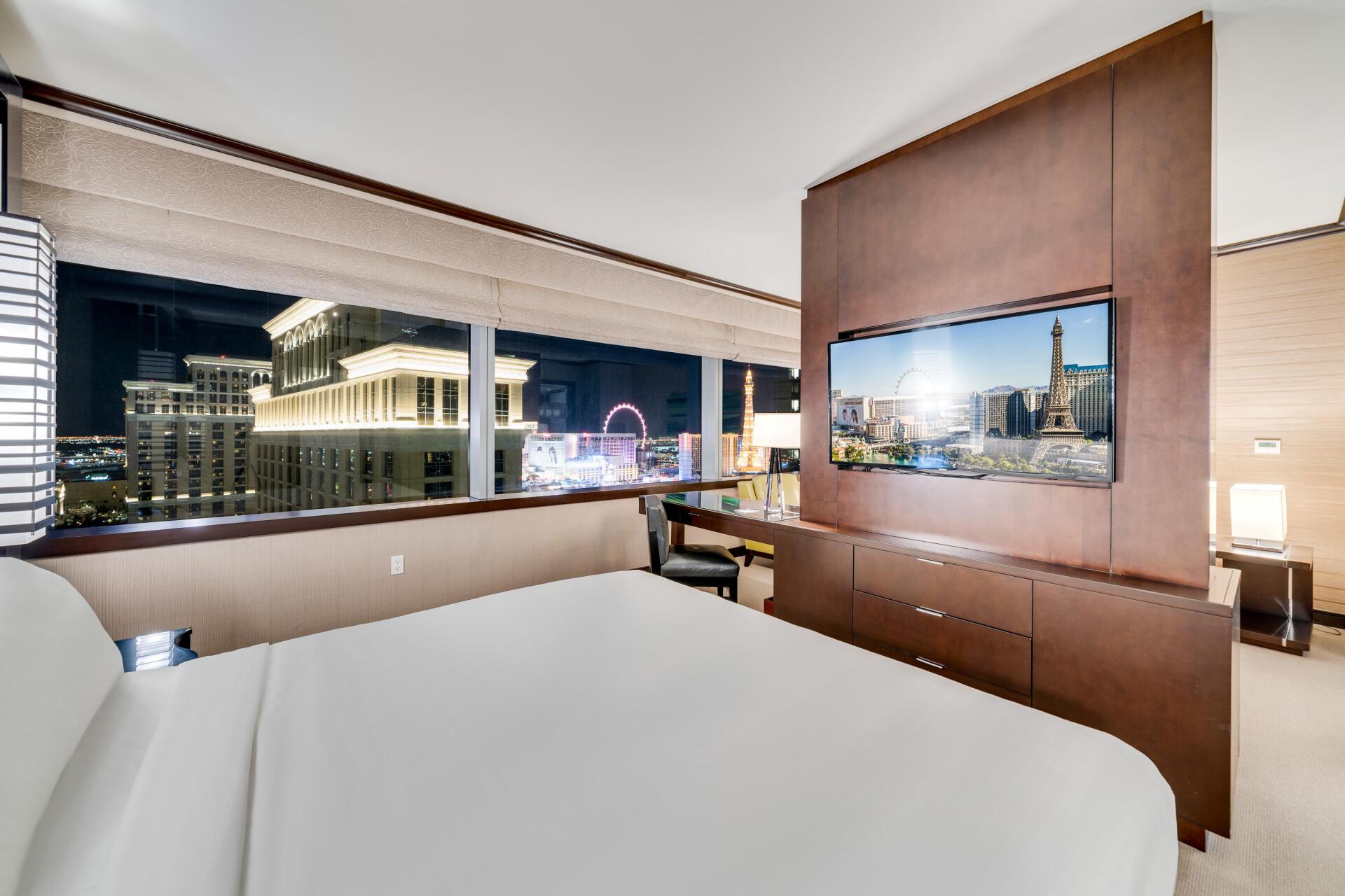 Hotel room with large bed, TV, and a nighttime city view through wide windows.