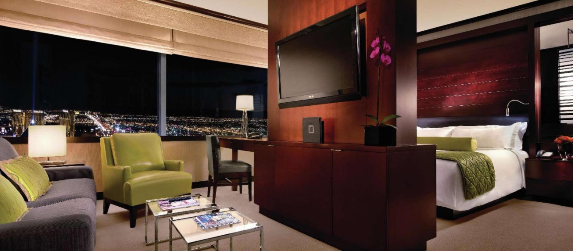 Modern hotel suite with city view, green chairs, a bed, and a wall-mounted TV in warm lighting