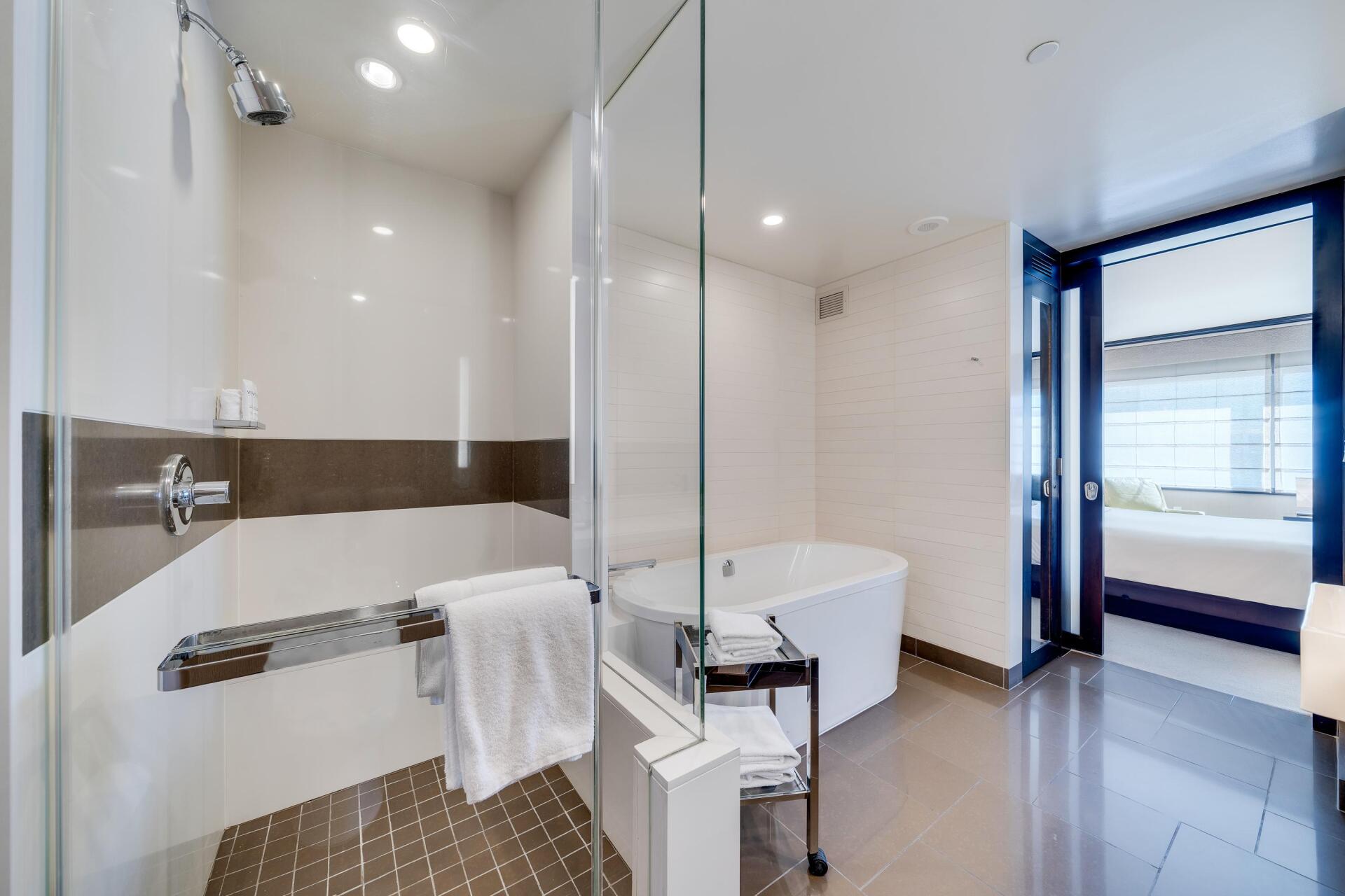 Modern bathroom with glass shower, white tub, towel rack, and open doorway to a bedroom