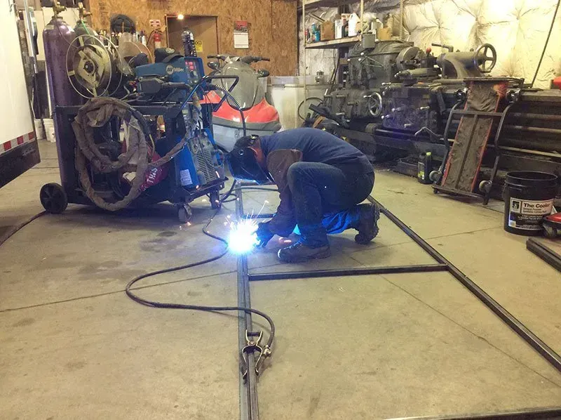 Person welding on the floor in a cluttered workshop, bright blue arc light illuminating the scene.
