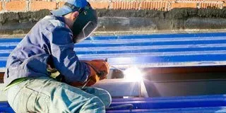 Worker welding metal on a blue structure outdoors, with bright sparks and a face shield