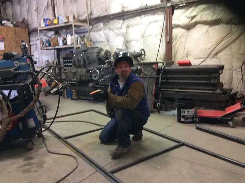 Person kneeling in a workshop, smiling beside a metal frame on the floor.