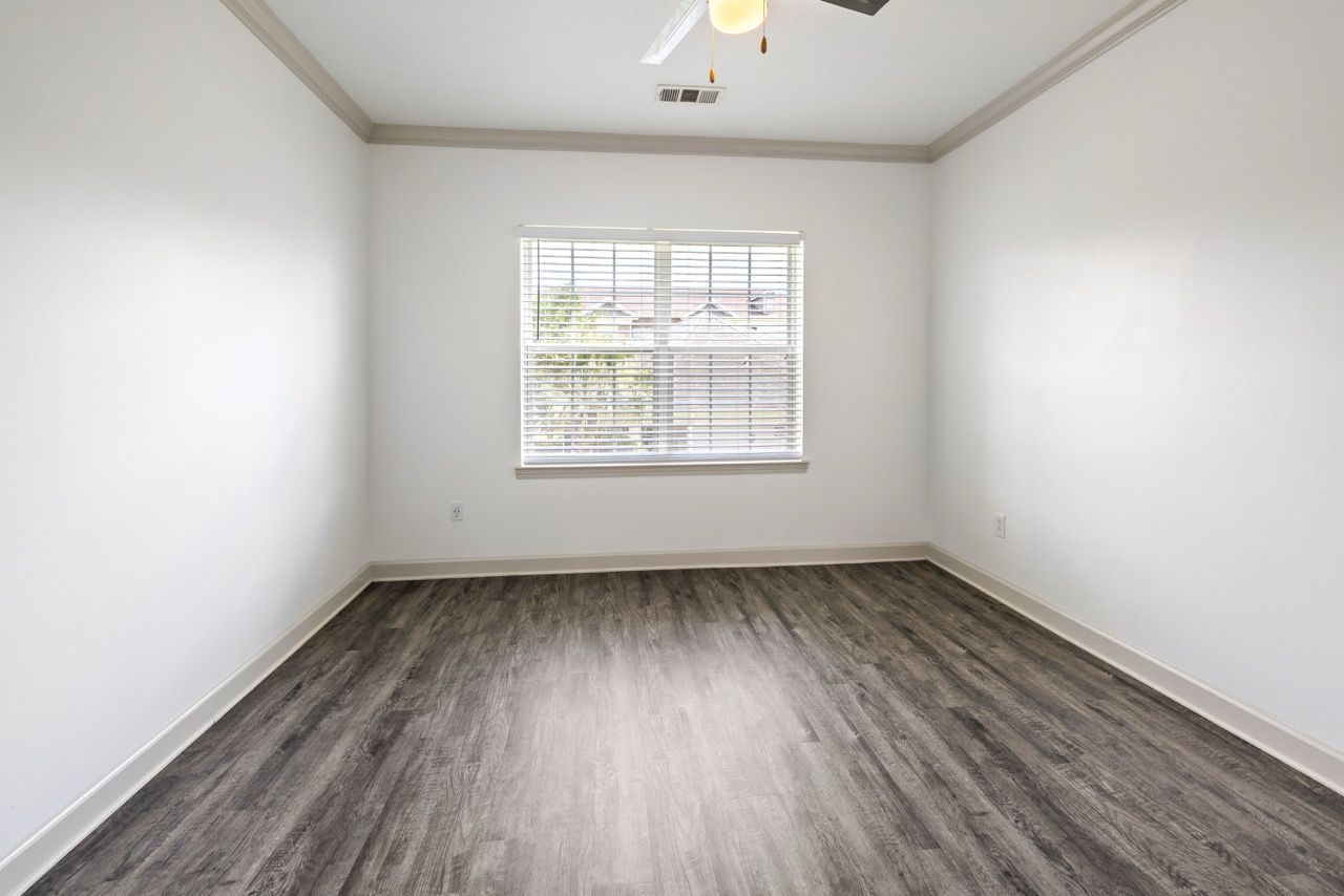 Empty bedroom with white walls, a window with blinds, and gray wood-look flooring.