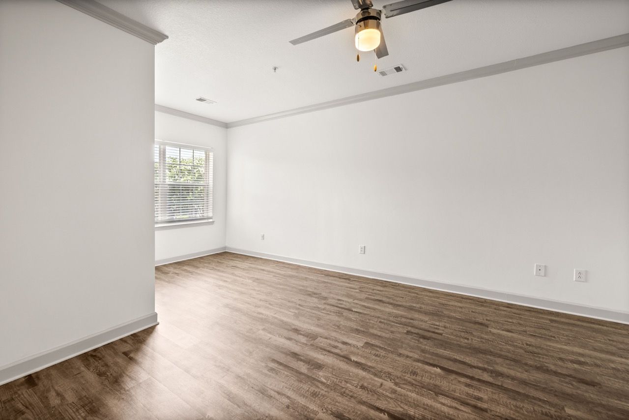 Empty living room with wood-look flooring, white walls, a ceiling fan, and a window with blinds.
