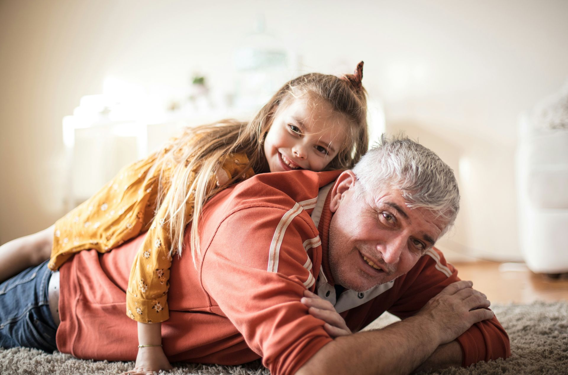 Smiling older man and child lying on a carpet, with the child hugging him from behind.