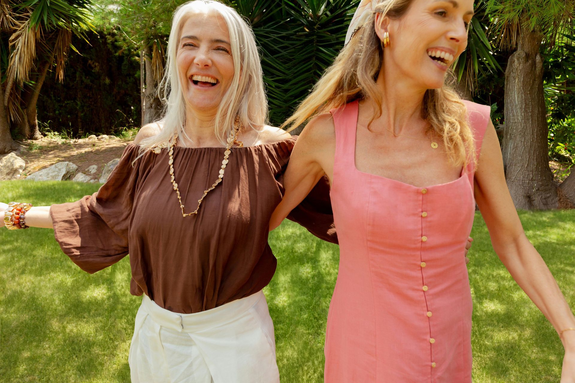 Two women smiling and walking arm-in-arm on a sunny lawn, wearing brown and pink dresses.