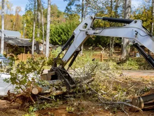 A white excavator arm clearing fallen tree branches and debris on a sunny, wooded residential property.