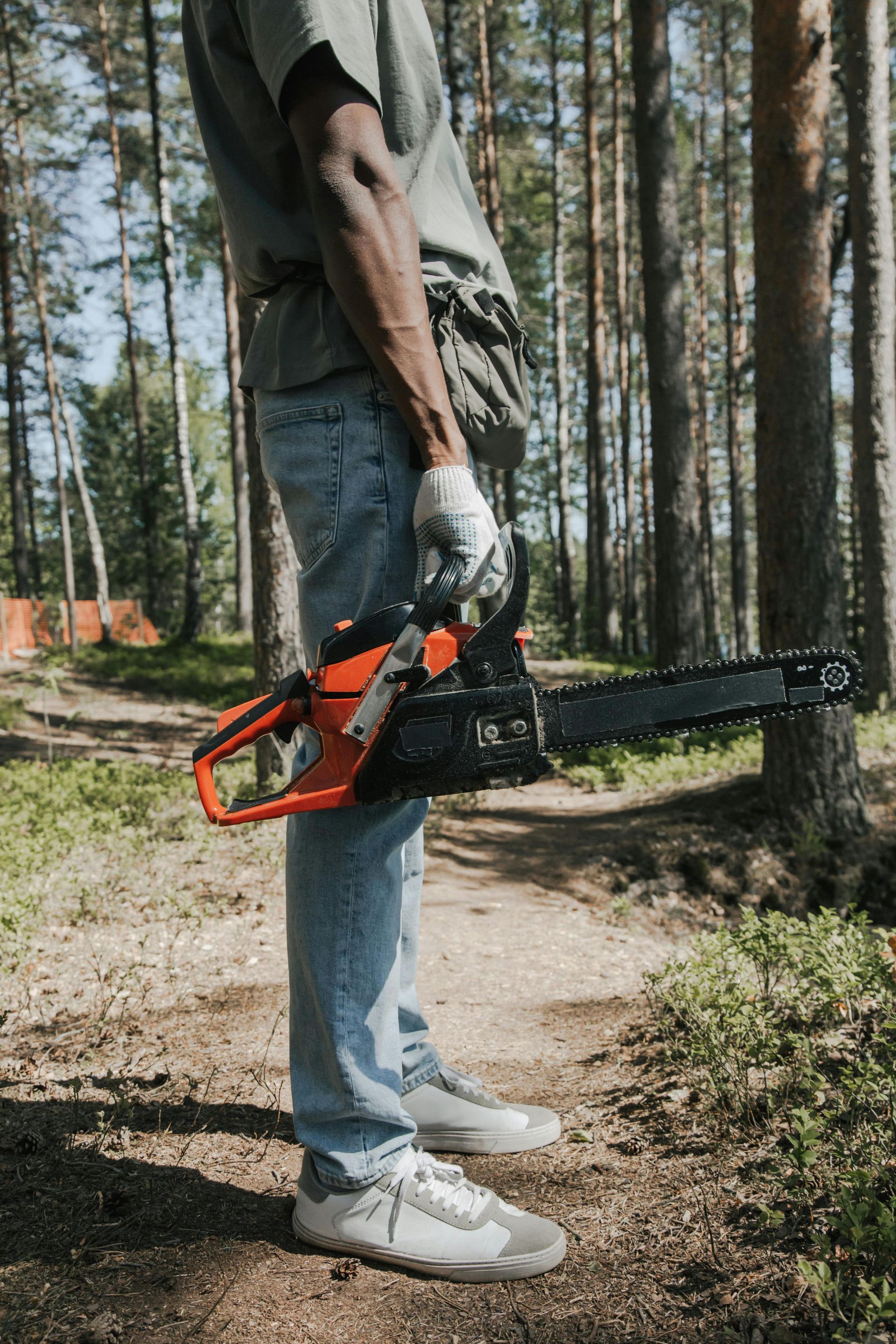 A person in casual clothes and gloves holds an orange chainsaw while standing on a forest path.