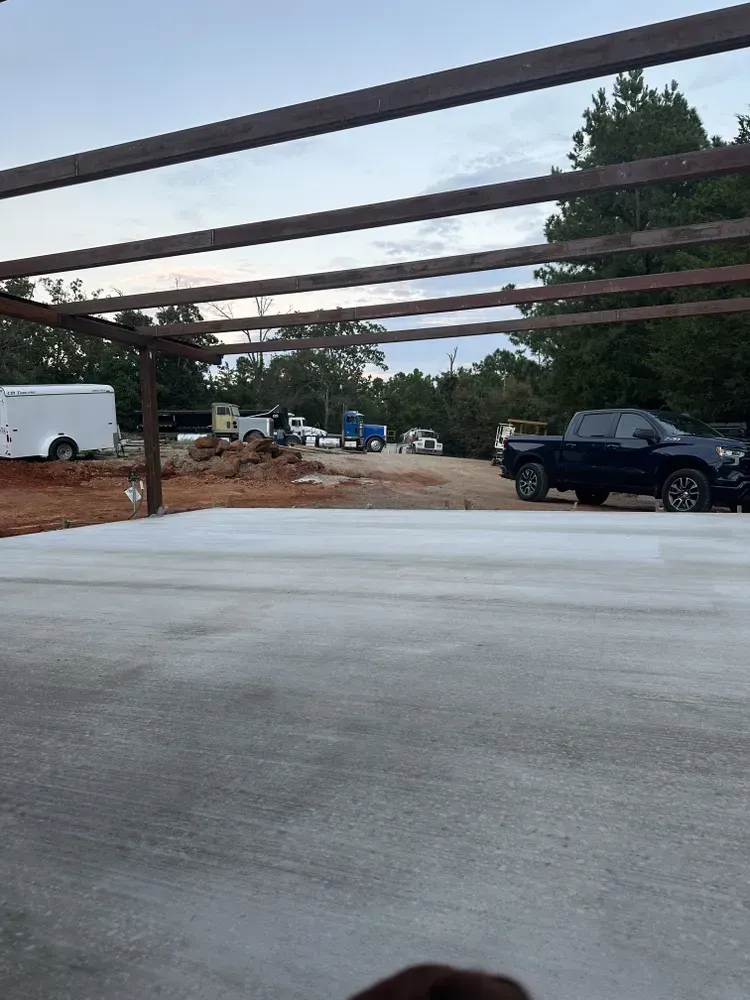 Concrete slab under construction with wooden beams overhead, parked truck, trailers, and trees in the background