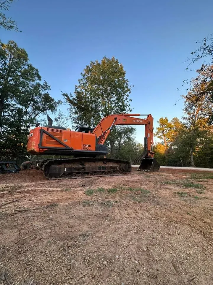 Orange excavator on tracks at a dirt lot with trees and blue sky.